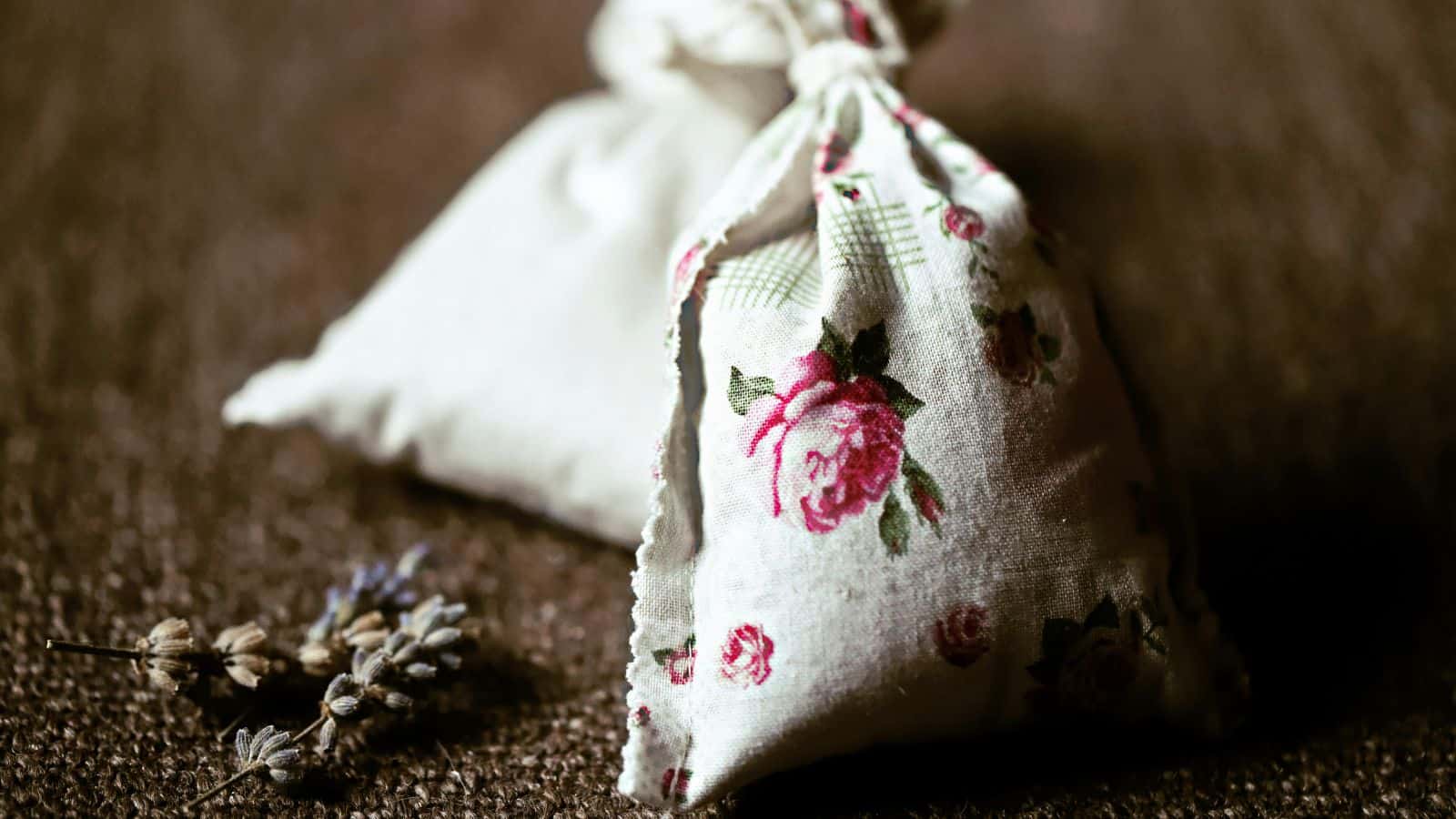 Two small fabric sachets sit on a textured surface. The front sachet features a floral design with pink roses, while the back sachet is plain. Dried lavender stems are placed beside them.