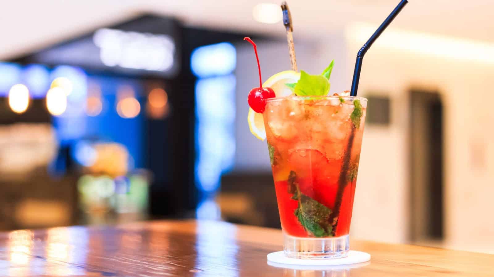 A refreshing red cocktail is served in a glass with ice, a lemon slice, mint leaves, a cherry, and a black straw. The glass is placed on a wooden surface, with blurred lights and a counter visible in the background.