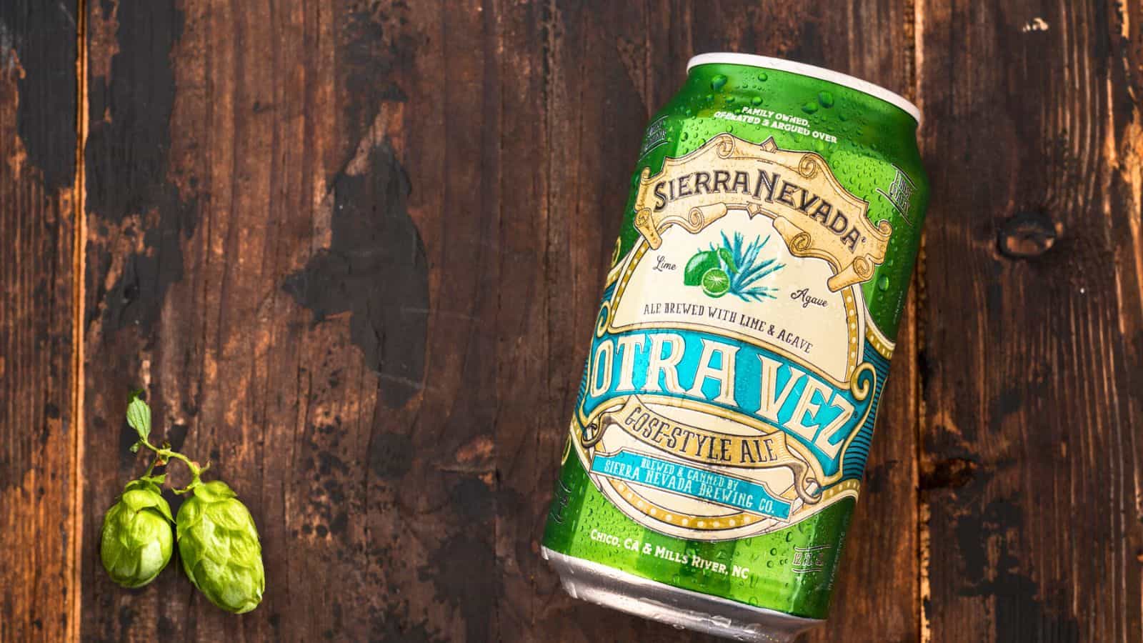 A can of Sierra Nevada Otra Vez beer is placed on a wooden surface, tilted slightly to the right. Next to the can, there are two green hops. The beer label displays the flavor as "gose-style ale brewed with lime & agave.
