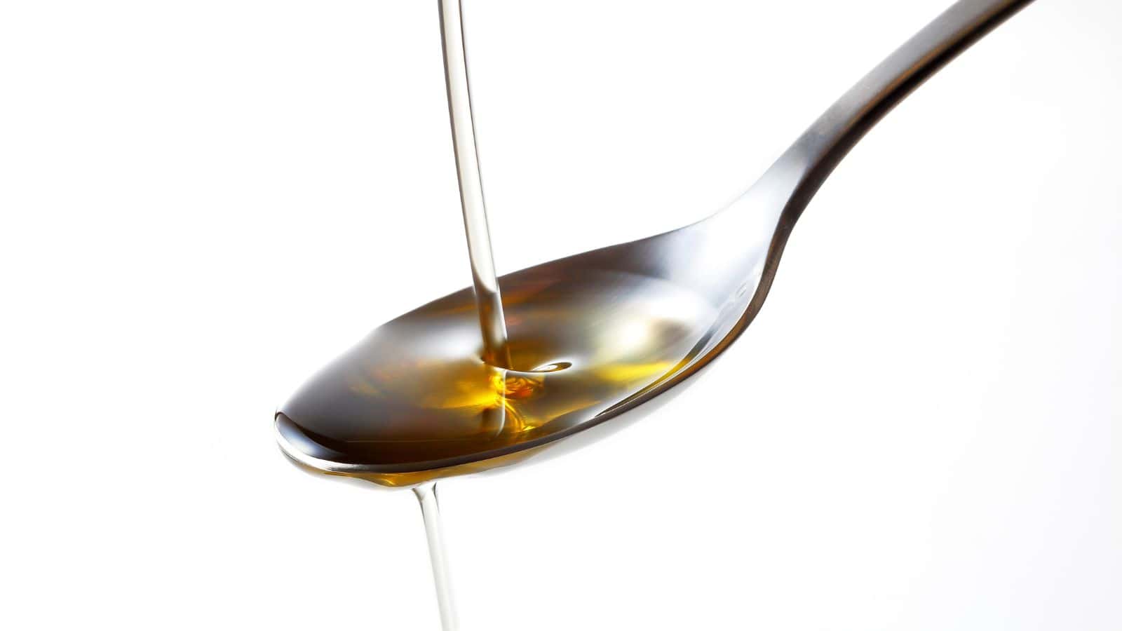 A close-up image of a metallic spoon with a stream of golden liquid, possibly oil or honey, being poured into it against a plain white background. The liquid is forming a small pool in the spoon.
