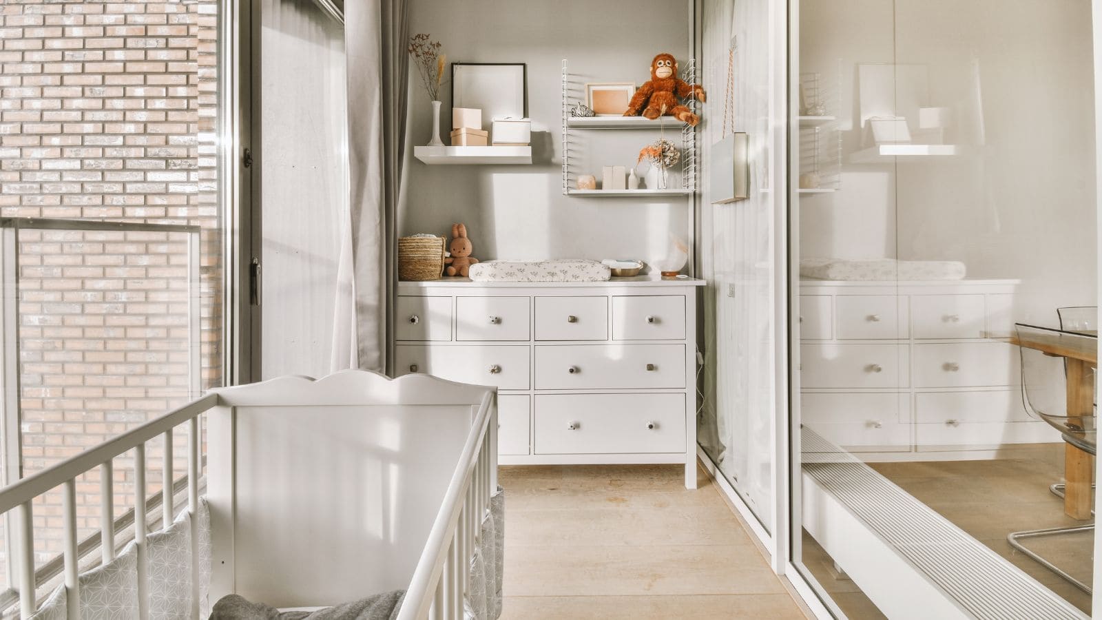 A nursery room with a white baby crib, a changing table with drawers, and shelves holding decorations, including a plush toy. The room has a large window with a brick wall outside and a mirrored closet. The floor is light wood.