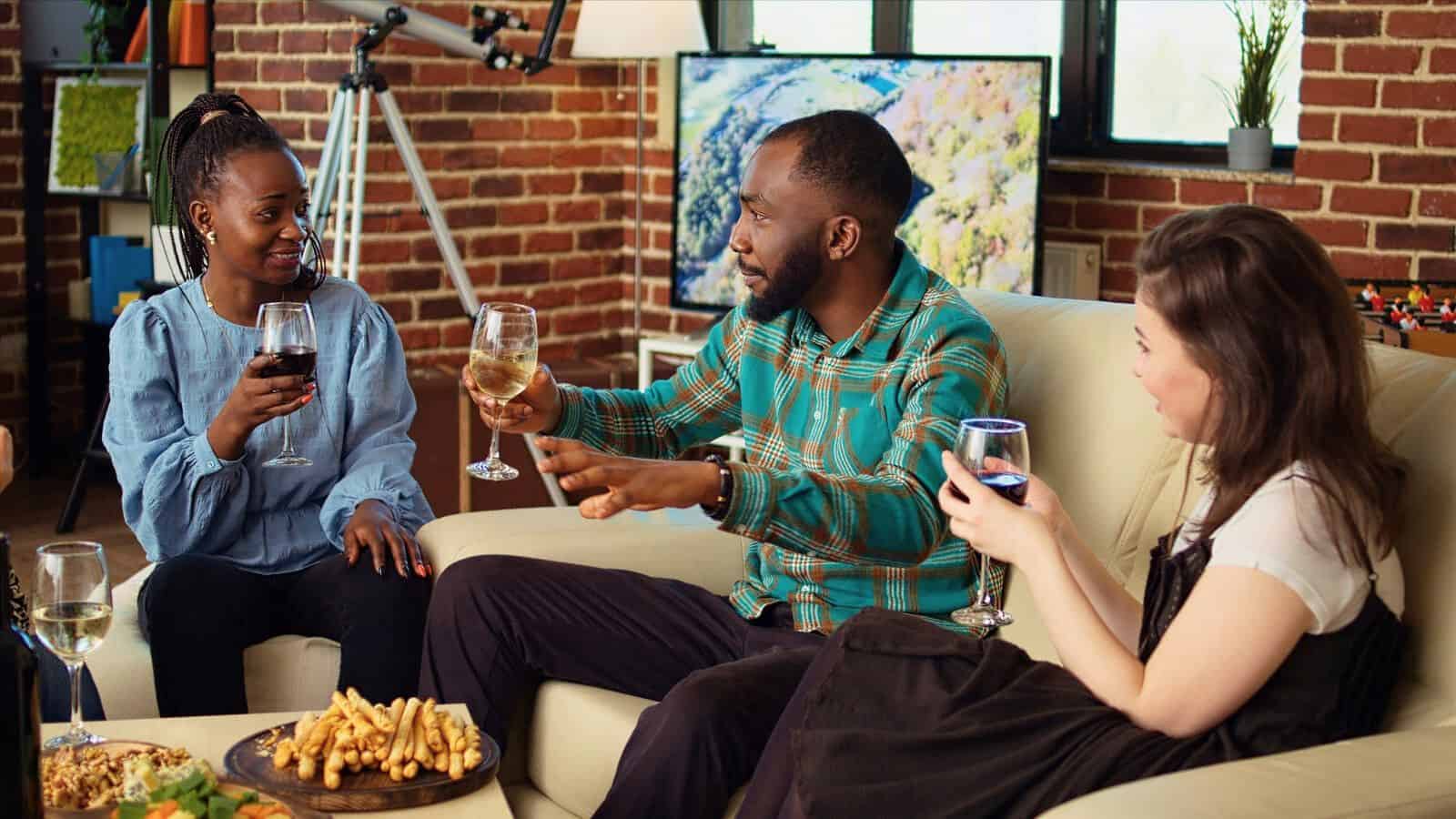 Three people sit on a sofa in a living room, each holding a glass of wine. The group is engaged in conversation. A table with snacks, including fries, is in front of them. A brick wall and telescope are visible in the background.
