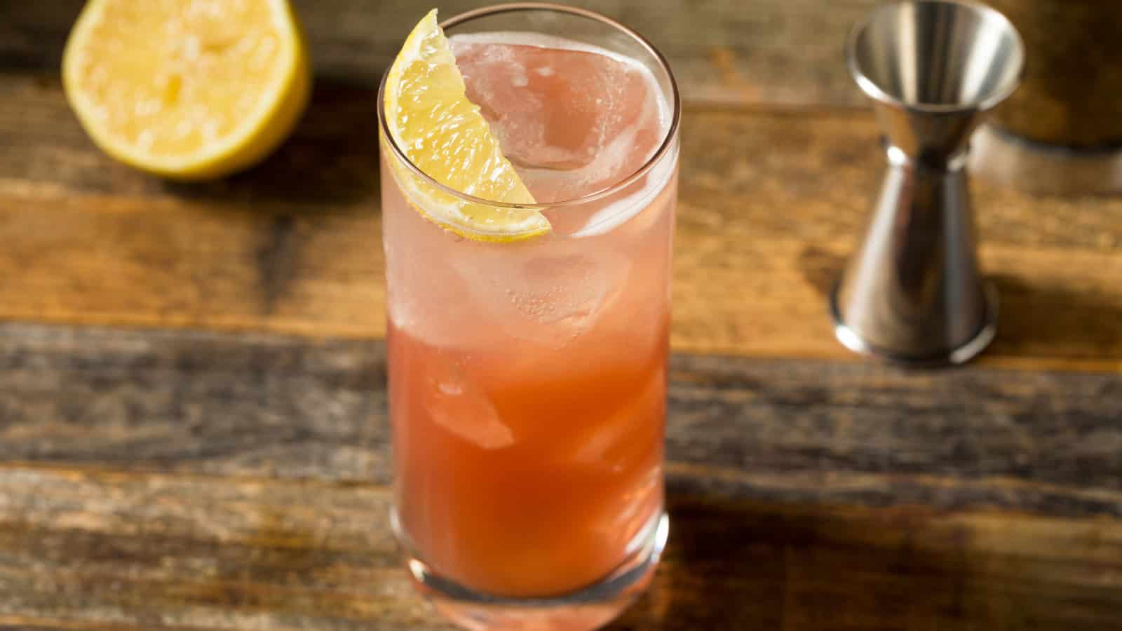 A tall glass filled with a pinkish cocktail is garnished with a lemon wedge. It sits on a wooden surface next to a metal jigger and a sliced lemon.