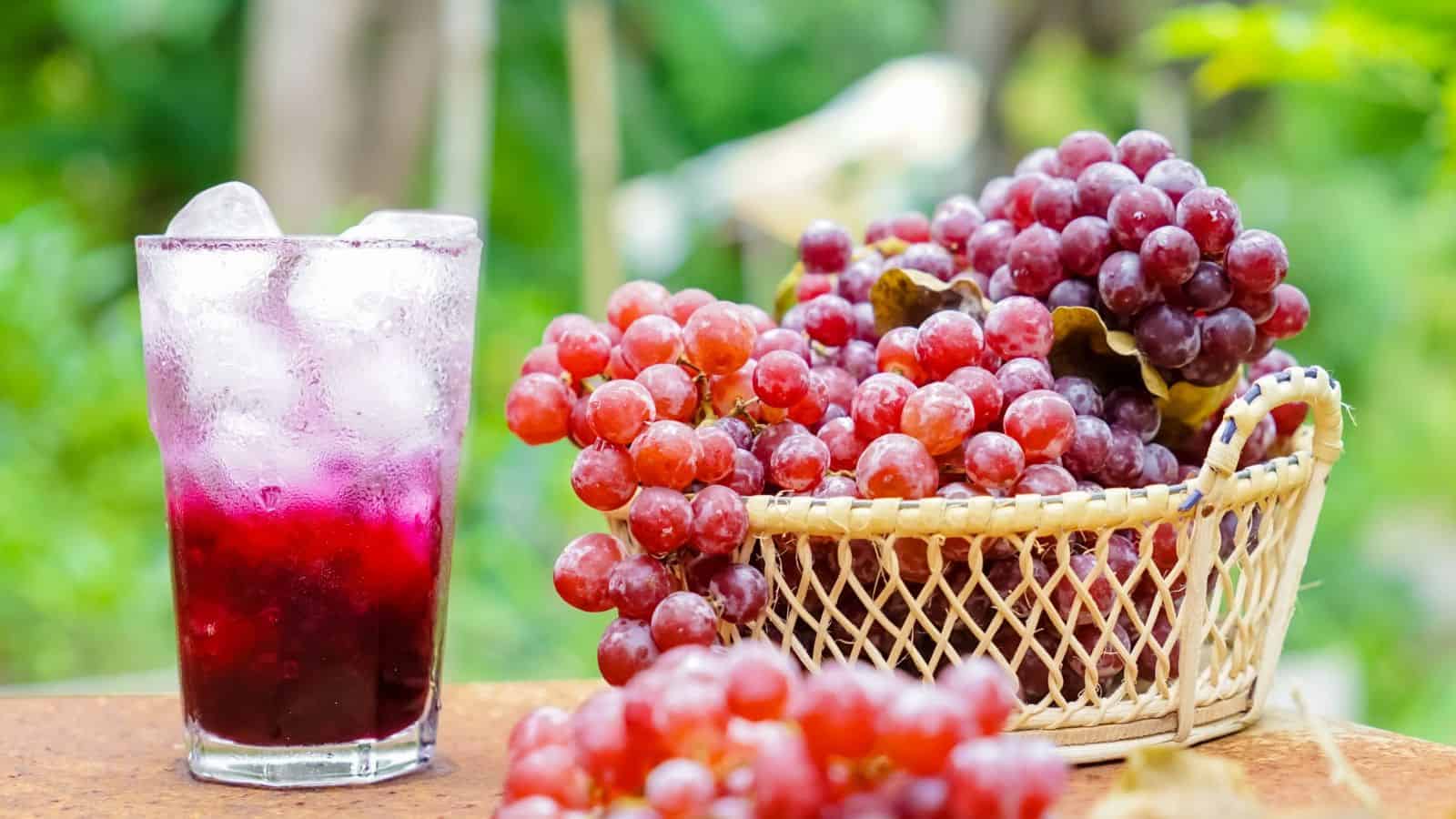 A glass of grape juice with ice is on a table beside a woven basket filled with red and purple grapes. The background is a blurred view of greenery.