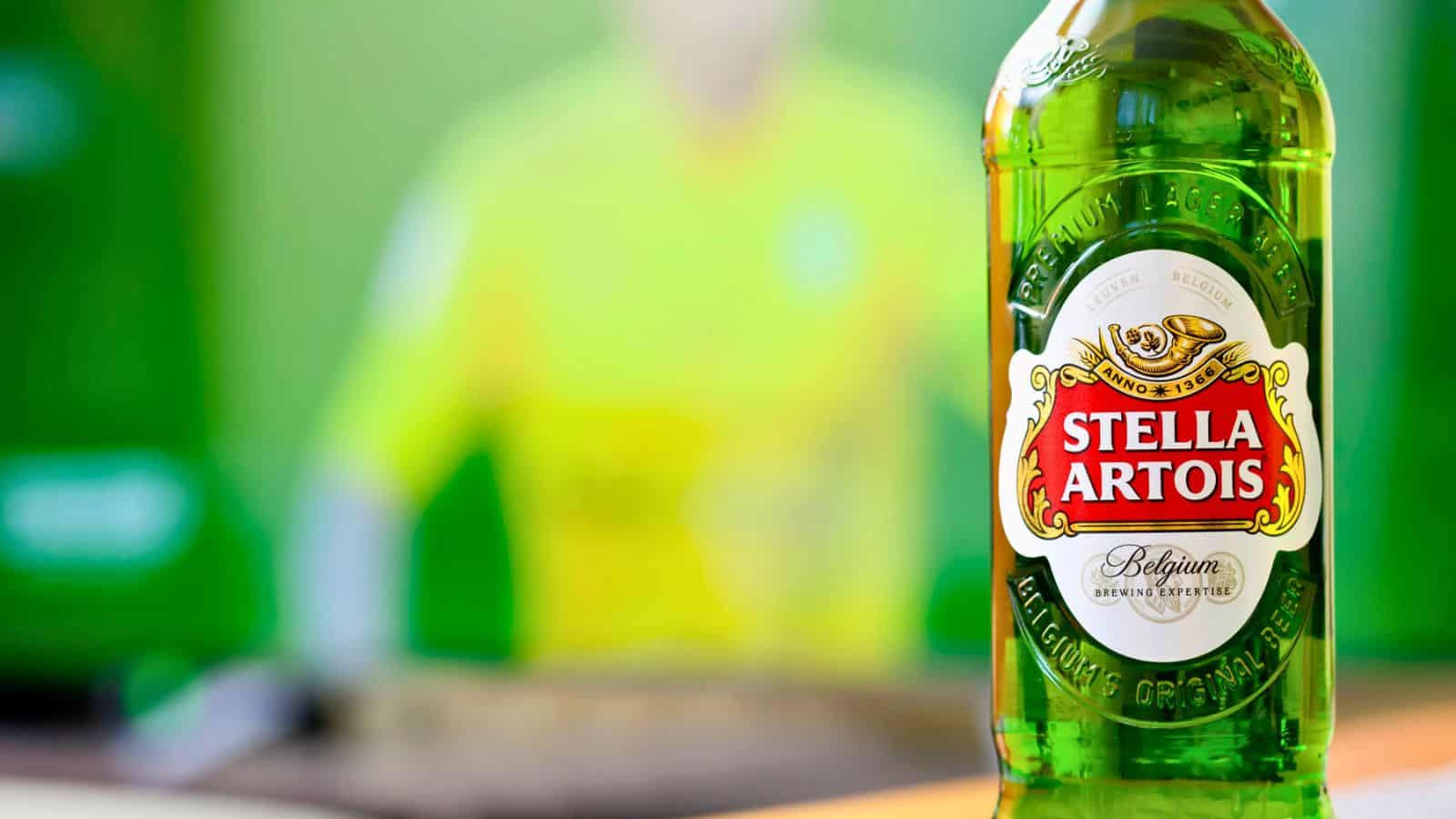 A close-up of a green glass beer bottle with a Stella Artois label. The background is blurred, featuring hints of green and yellow tones.