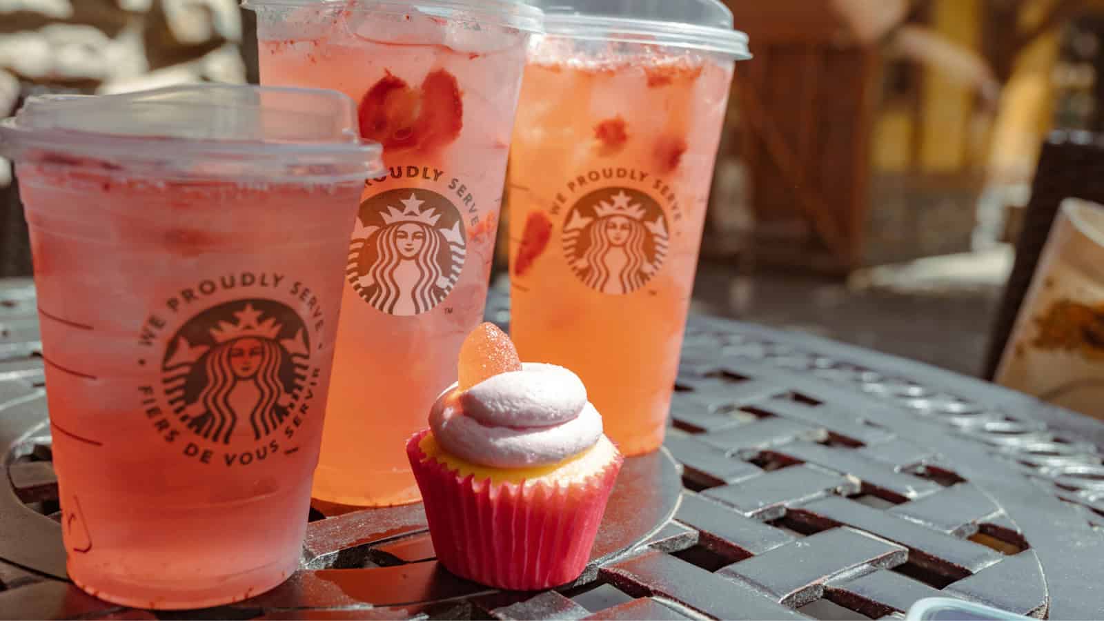 Three iced pink drinks in Starbucks cups are on a metal table, along with a cupcake topped with pink and white frosting. The background shows a sunlit outdoor setting.