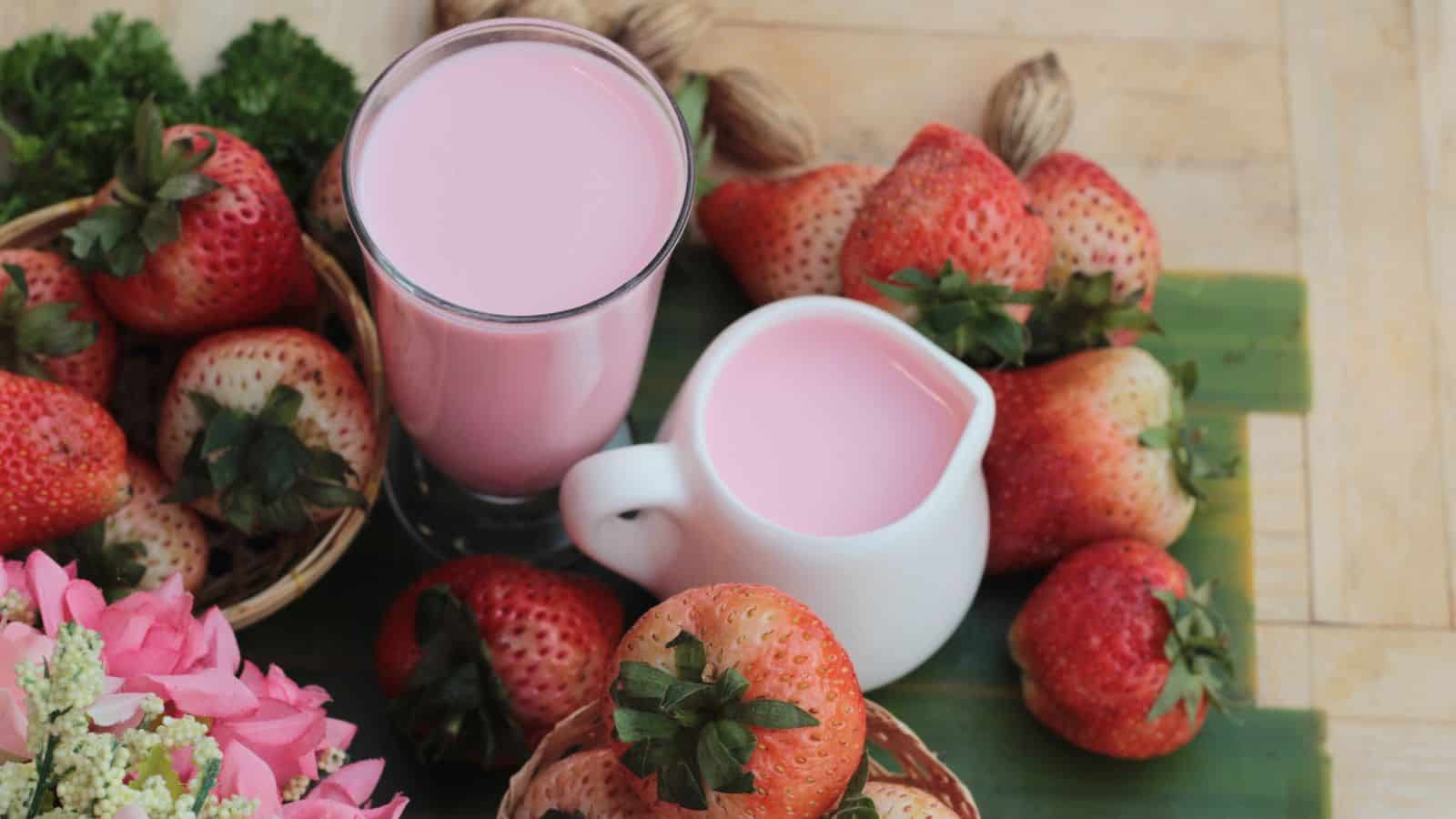 A glass of pink strawberry milk and a small white pitcher with more milk are surrounded by fresh strawberries on a wooden surface. Pink flowers and green leaves are placed nearby for decoration.