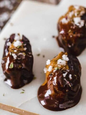 Three dates covered in dark chocolate and sprinkled with sea salt and herbs are arranged on a white cutting board. Each of these stuffed dates with almond butter exudes a touch of decadence. The board is placed on a wooden surface, with scattered salt flakes visible around the dates.