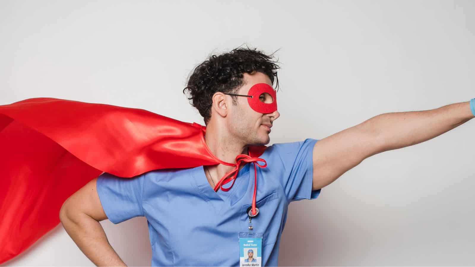 A person in blue scrubs with a name tag poses heroically, wearing a red cape and red mask, against a plain white background. They are extending one arm forward as if flying.