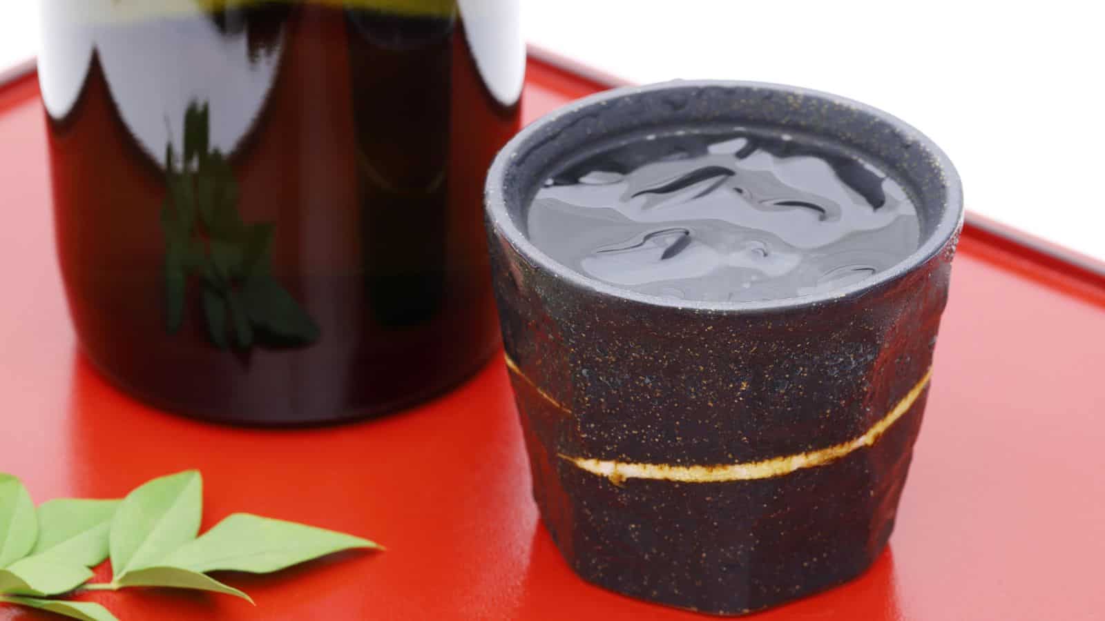 A rustic ceramic cup filled with a dark liquid is placed on a red surface next to a bottle. There are green leaves nearby, adding a touch of nature to the scene.