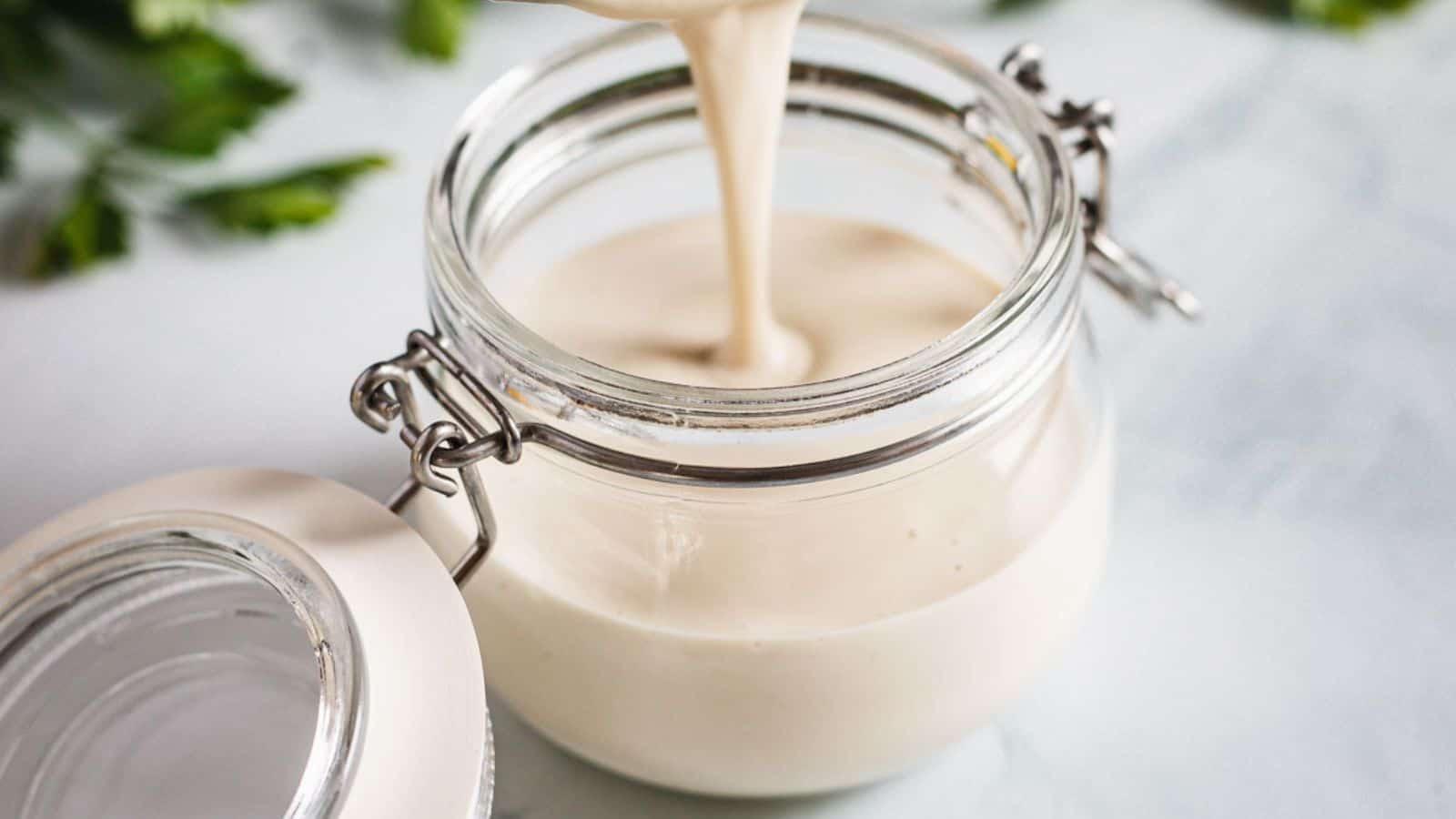 A thick, creamy liquid is being poured into an open glass jar with a metal clasp lid. The jar is placed on a light-colored surface, with greenery blurred in the background.