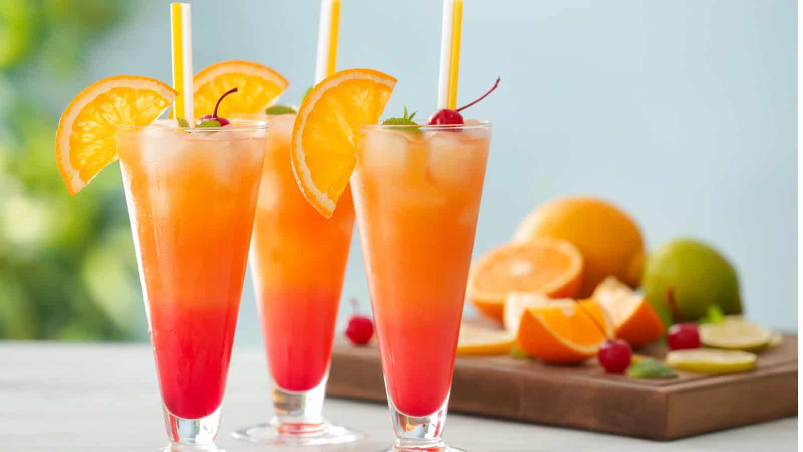 Three tropical cocktails in tall glasses, featuring a gradient from red to orange, garnished with orange slices and cherries, with yellow straws. In the background, sliced oranges and limes are on a wooden board.