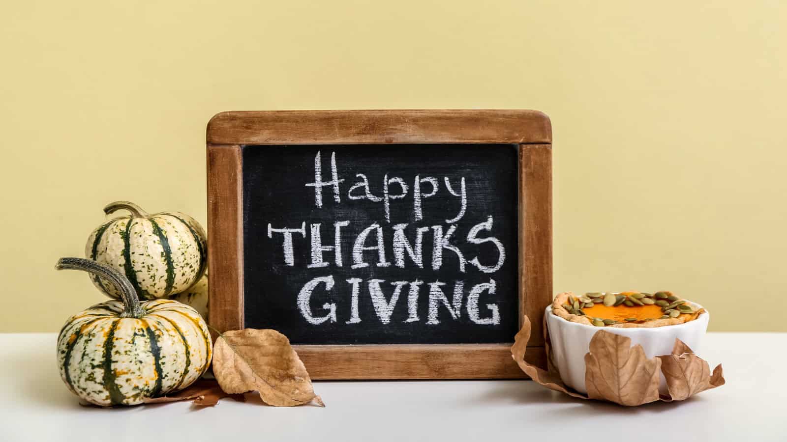 A small blackboard with "Happy Thanksgiving" written on it is placed on a white surface. Next to the board are two small decorative pumpkins and a white dish with orange content, surrounded by dried leaves. The background is a pale yellow.