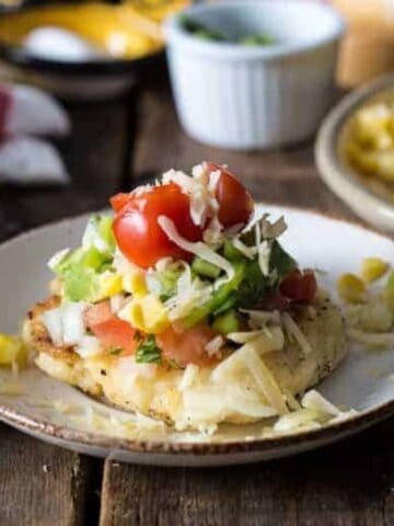 A plate with grilled chicken breast topped with chopped avocado, tomatoes, onions, and shredded cheese. In the background, there's a dish of corn and other small bowls containing green onions and herbs on a wooden table.