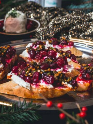 A festive platter features toasted bread topped with cream cheese, roasted cranberries, and a relish-like topping. The plate is decorated with dark, glittery fabric, surrounded by greenery and red berries, creating a holiday atmosphere.
