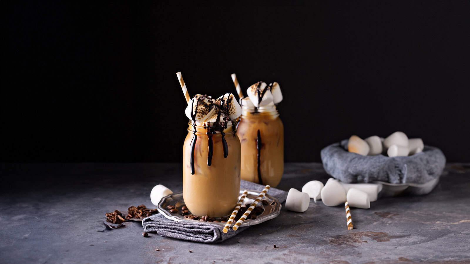 Two mason jars filled with a creamy coffee drink topped with whipped cream, chocolate drizzle, and marshmallows. Striped straws are inserted into the drinks. A few marshmallows and chocolate pieces are scattered around on a dark surface.