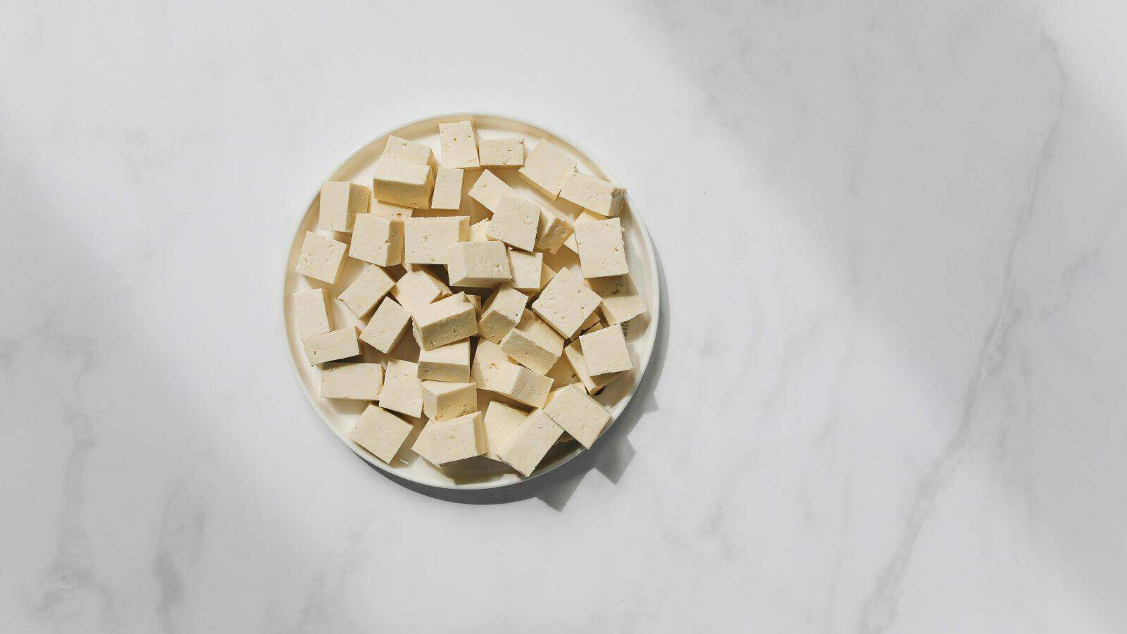 A round white plate is filled with small cubes of tofu on a light marble surface. The tofu is arranged neatly, casting subtle shadows. The scene is evenly lit, highlighting the creamy texture of the tofu against the smooth background.