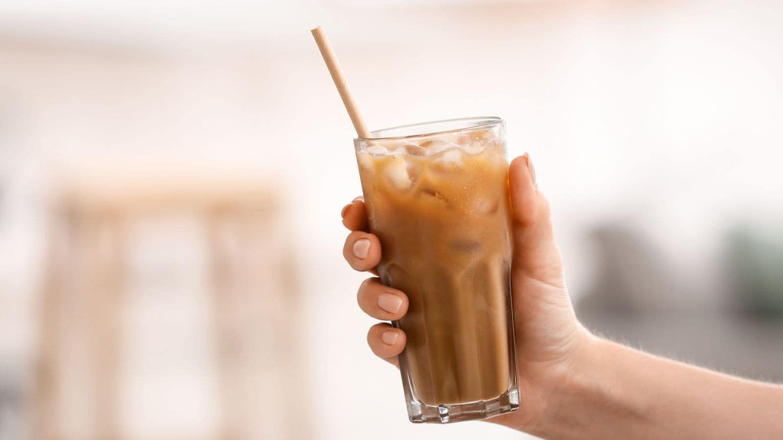 A hand holding a tall glass of iced coffee with a straw. The drink is filled with ice cubes and has a creamy appearance. The background is softly blurred.
