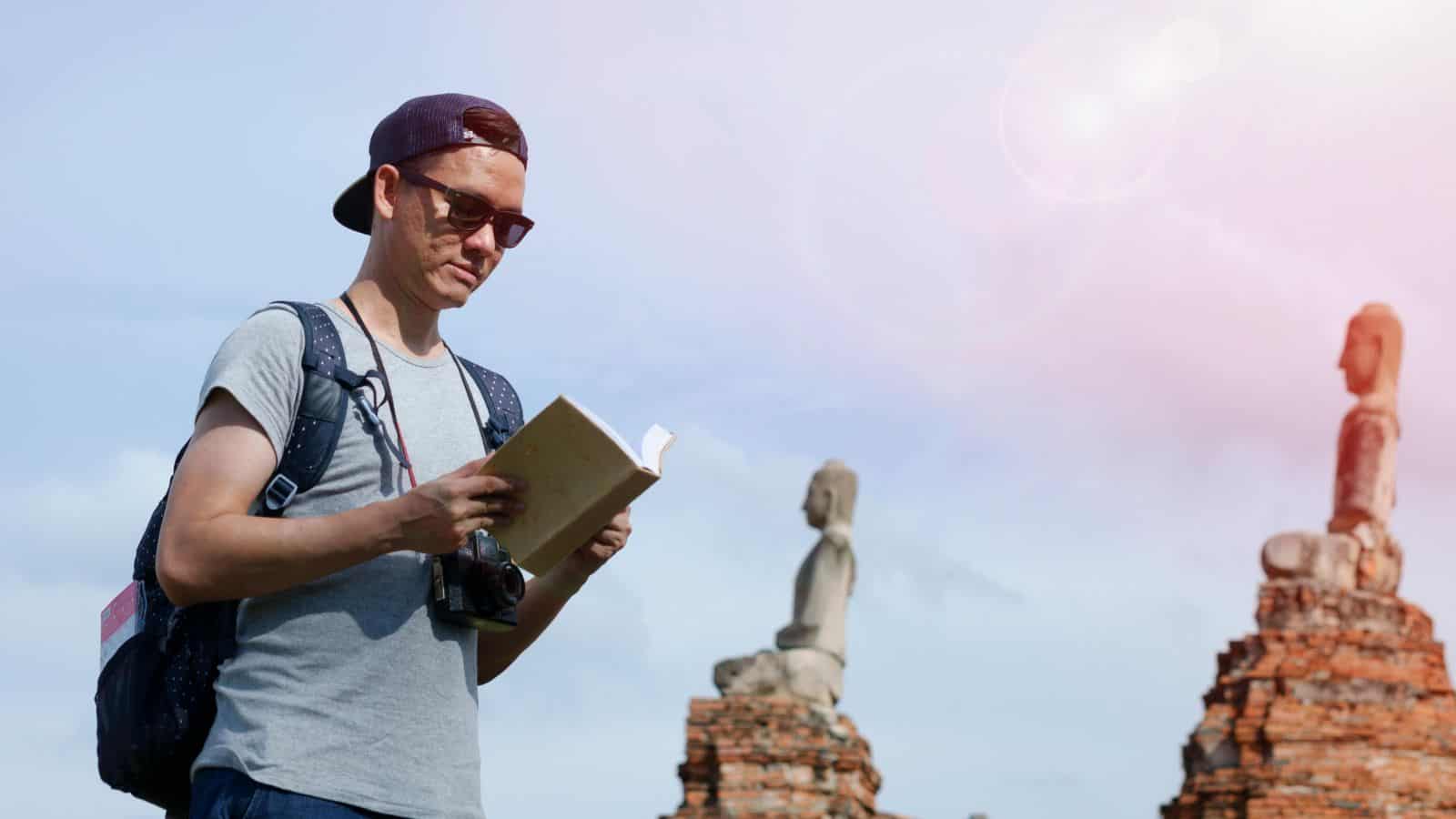 A person with a backpack and camera, wearing sunglasses and a cap, stands outdoors reading a book. In the background, there are ancient stone statues and a clear sky.