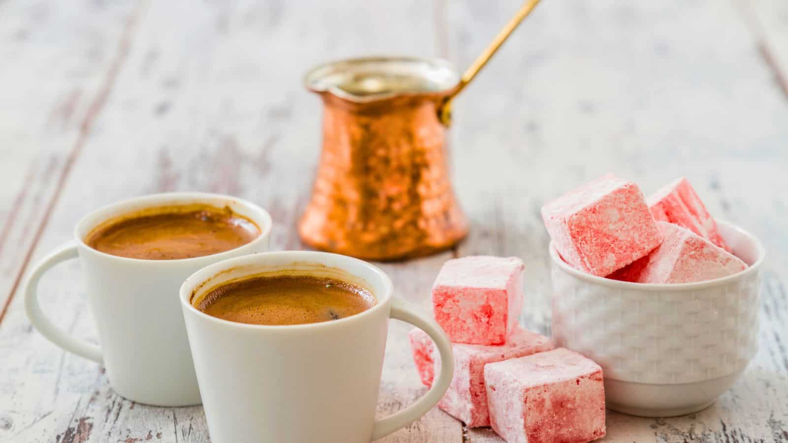 Two white cups filled with Turkish coffee sit on a wooden surface. A copper cezve is in the background. A bowl beside the cups contains pink and white Turkish delight pieces.