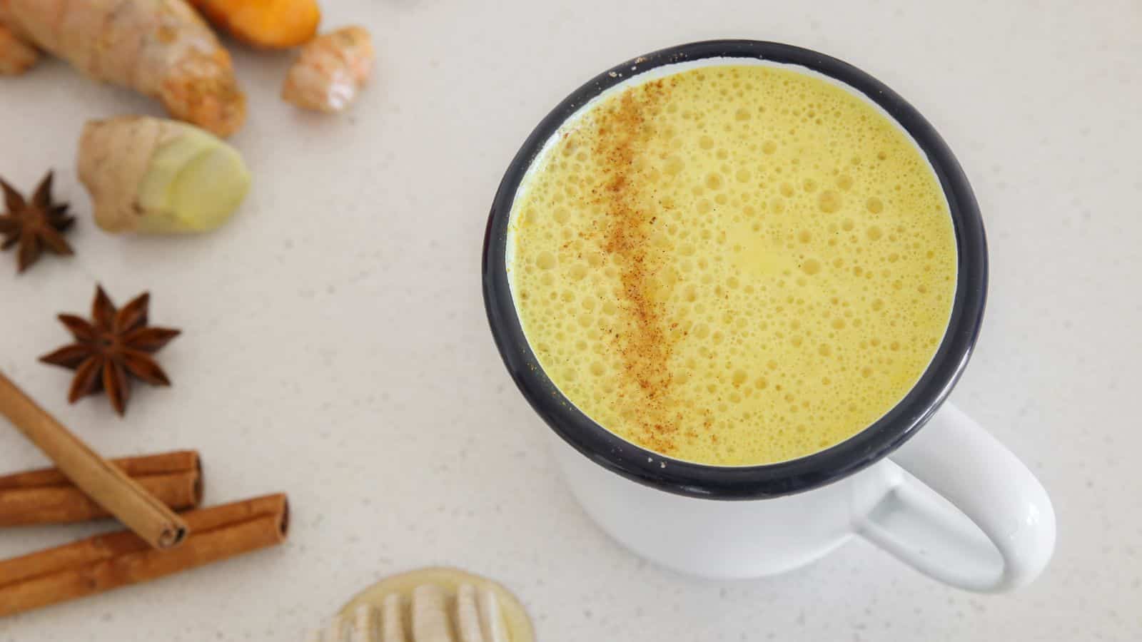 A white mug filled with yellow turmeric latte sprinkled with cinnamon on top, placed on a white countertop. Surrounding the mug are pieces of ginger, turmeric, star anise, and cinnamon sticks.