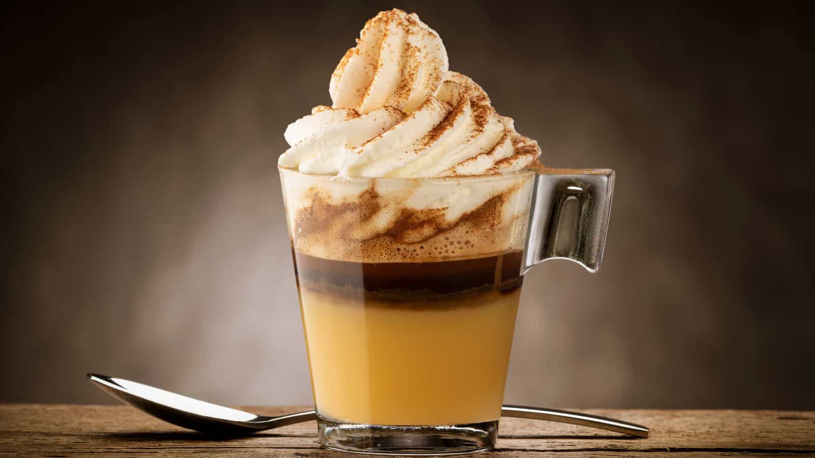 A glass mug filled with layered coffee and cream sits on a wooden surface, reminiscent of underrated Starbucks flavors. The coffee is topped with whipped cream sprinkled with cocoa powder, while a metal spoon rests beside it. The background is softly lit with a warm tone.