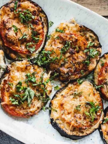 A white plate holds several slices of grilled eggplant topped with melted cheese, tomato slices, and fresh herbs. The plate is set on a wooden surface, partially covering a dark cloth napkin.