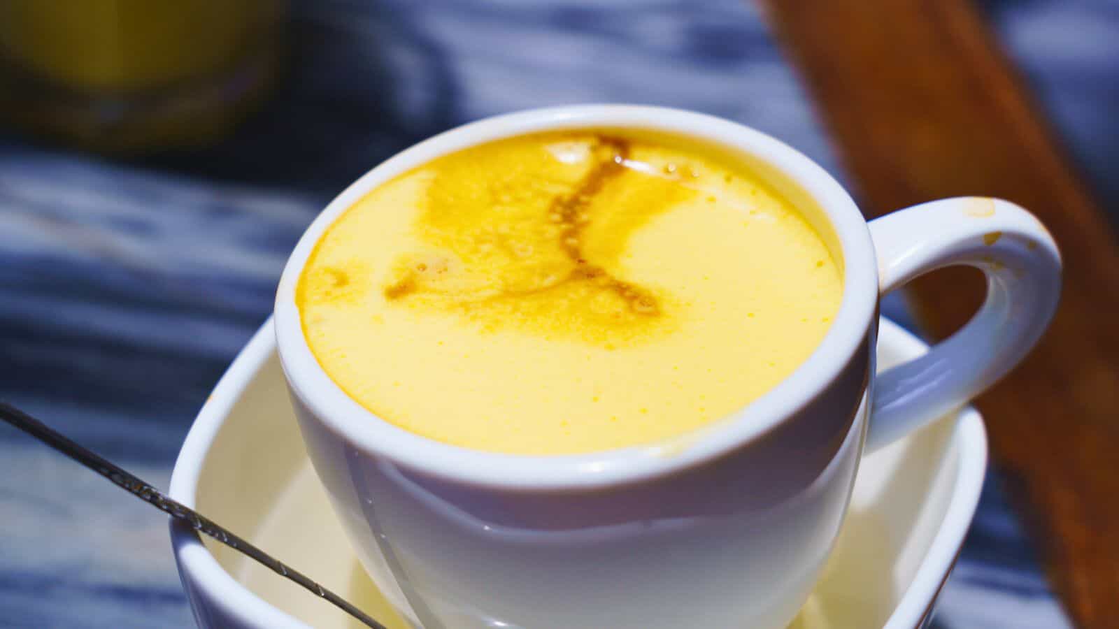 A white ceramic cup filled with a creamy, golden-yellow beverage, likely a latte or similar drink, placed on a matching saucer. A silver spoon rests beside the cup on a stone-textured surface.