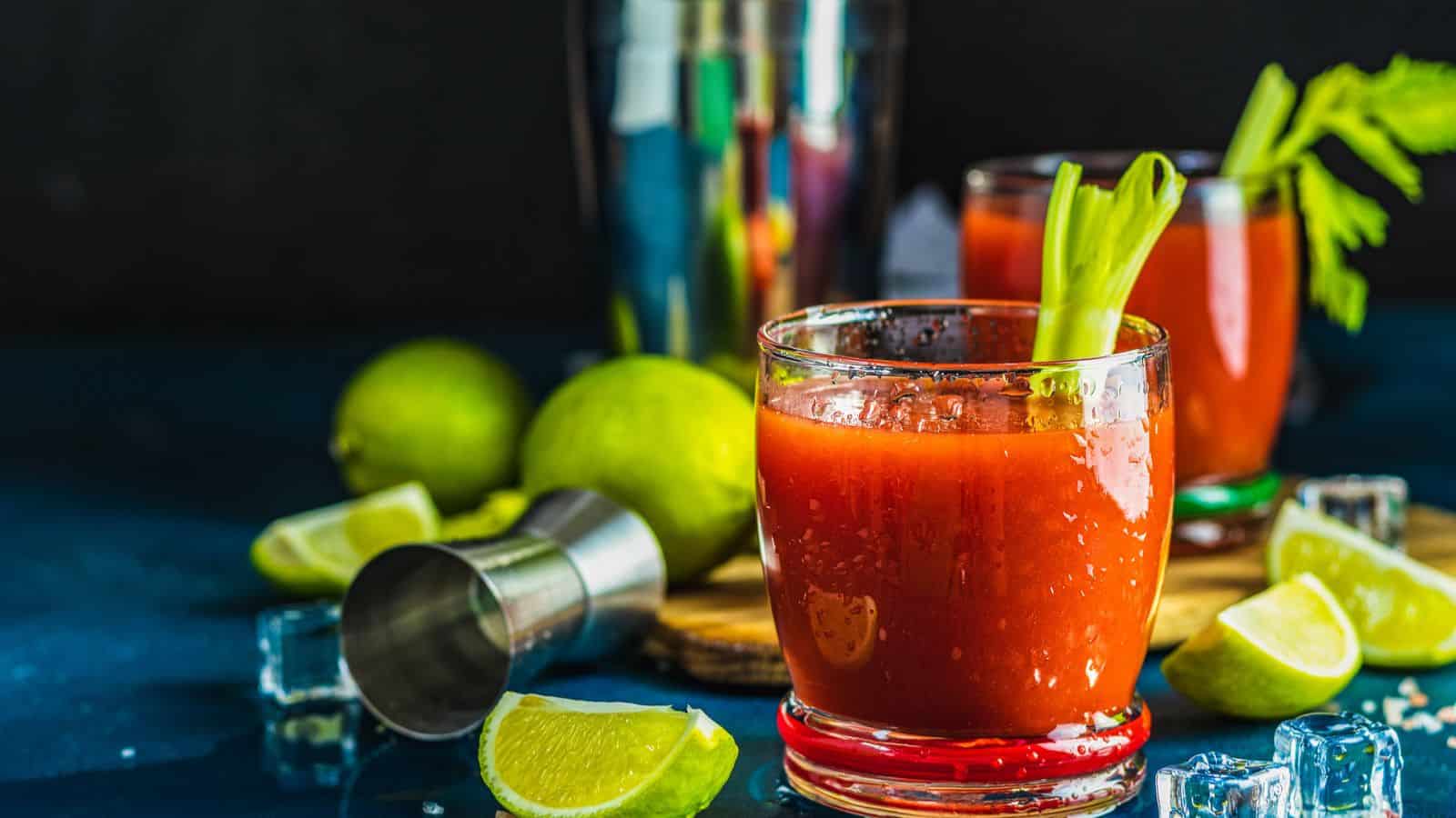 A close-up of two Bloody Mary cocktails in clear glasses, garnished with celery sticks. Lime wedges, a metal shaker, and ice cubes are scattered around on a dark surface. The background is blurred.