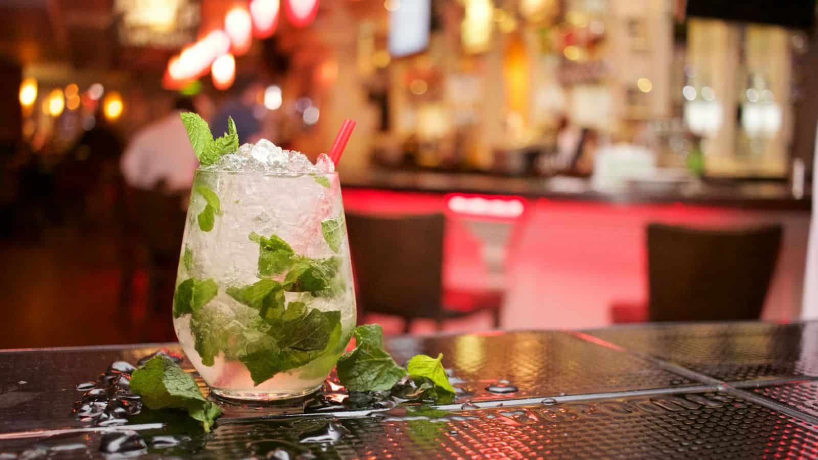 A cocktail glass filled with a mojito, featuring crushed ice, mint leaves, and a lime wedge, sits on a bar counter. The background shows a dimly lit bar with red lighting and blurry bar stools.
