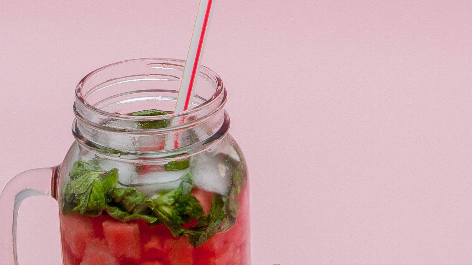 A mason jar filled with a red beverage, garnished with mint leaves and ice, sits on a pink surface. A red and white striped straw is placed inside the jar.