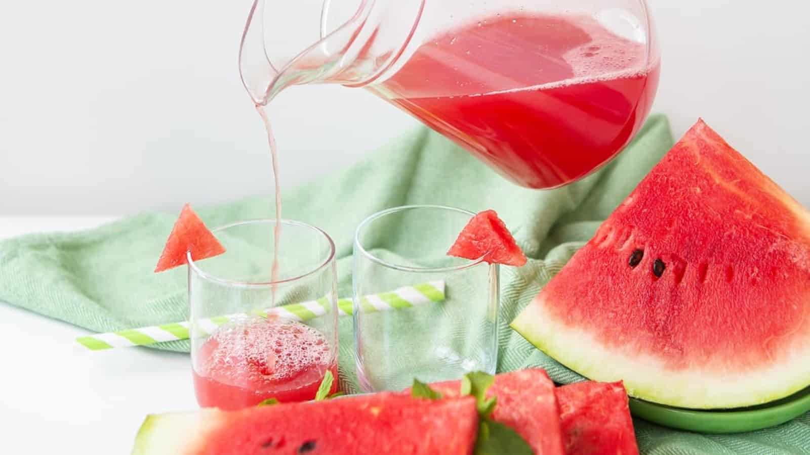A glass pitcher pours watermelon juice into a glass. Two empty glasses with green straws stand nearby. A green cloth lies beneath. Several slices of watermelon are arranged on a white surface.