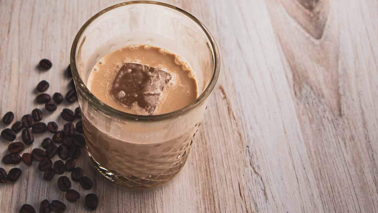 A glass of iced coffee with a single ice cube sits on a wooden surface, surrounded by scattered coffee beans.