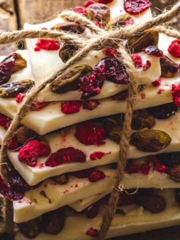 A stack of white chocolate bark pieces tied with twine rests on a wooden surface. The bark is topped with dried cranberries, raspberries, and nuts, providing a colorful and textured appearance.