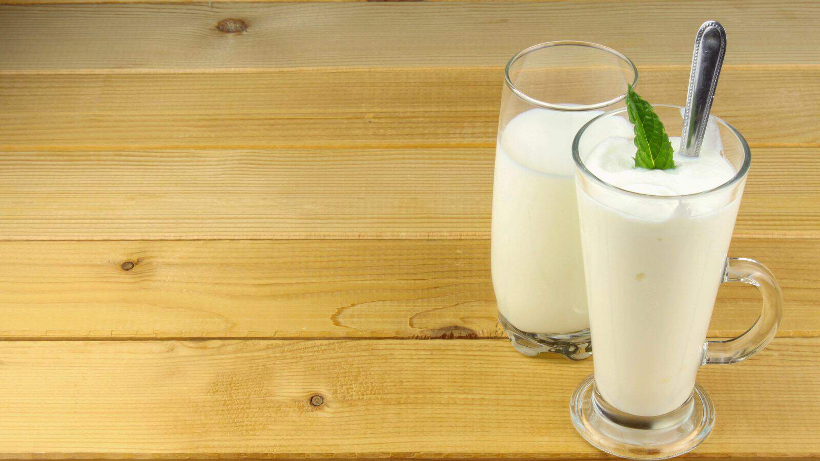 Two glasses of a creamy white beverage on a wooden surface. One glass is garnished with a mint leaf and a spoon, while the other is plain.