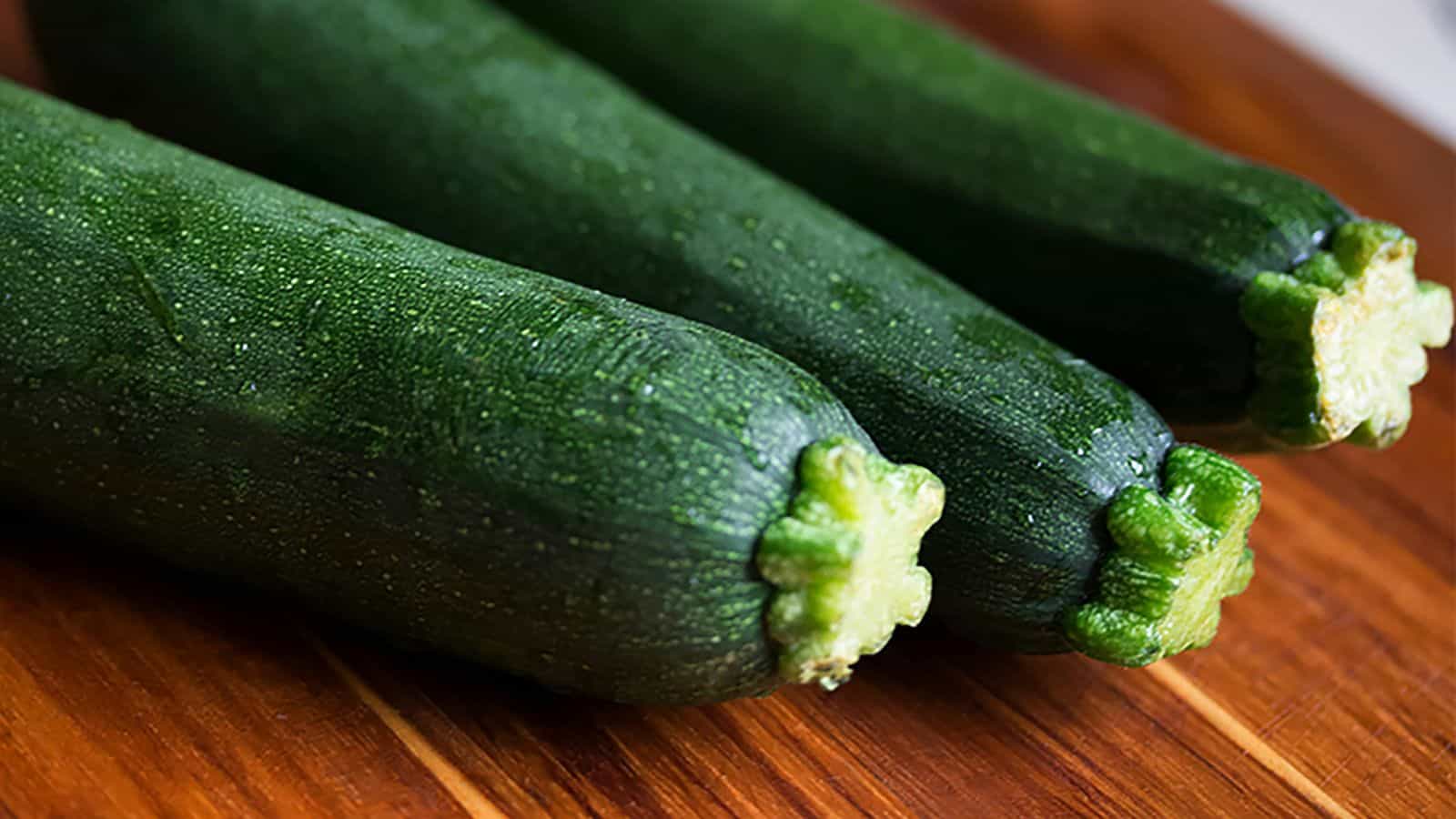 Three whole zucchinis are placed side by side on a wooden surface. The zucchinis are dark green with a smooth texture.