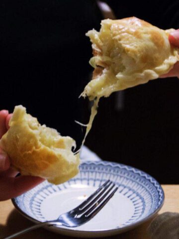 A person holds a piece of bread with melted cheese stretching between two halves over a small plate on a wooden table. A silver fork is placed next to the plate.