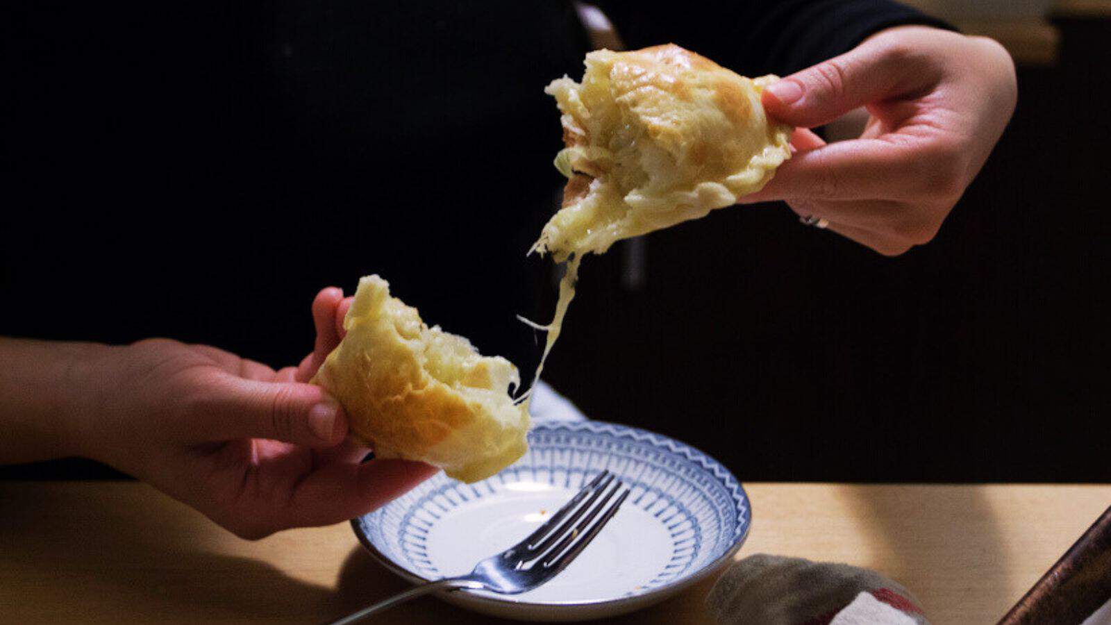 A person holds a piece of bread with melted cheese stretching between two halves over a small plate on a wooden table. A silver fork is placed next to the plate.