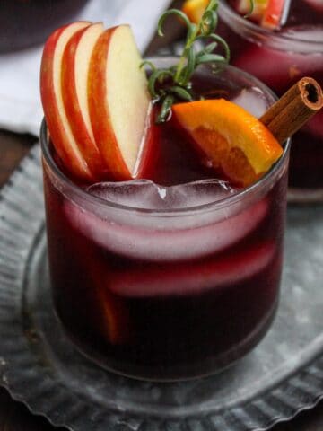 Two glasses of dark red sangria sit on a metal tray. Each glass is garnished with apple slices, orange wedges, a cinnamon stick, and a sprig of rosemary. A small plate with more orange slices is nearby.
