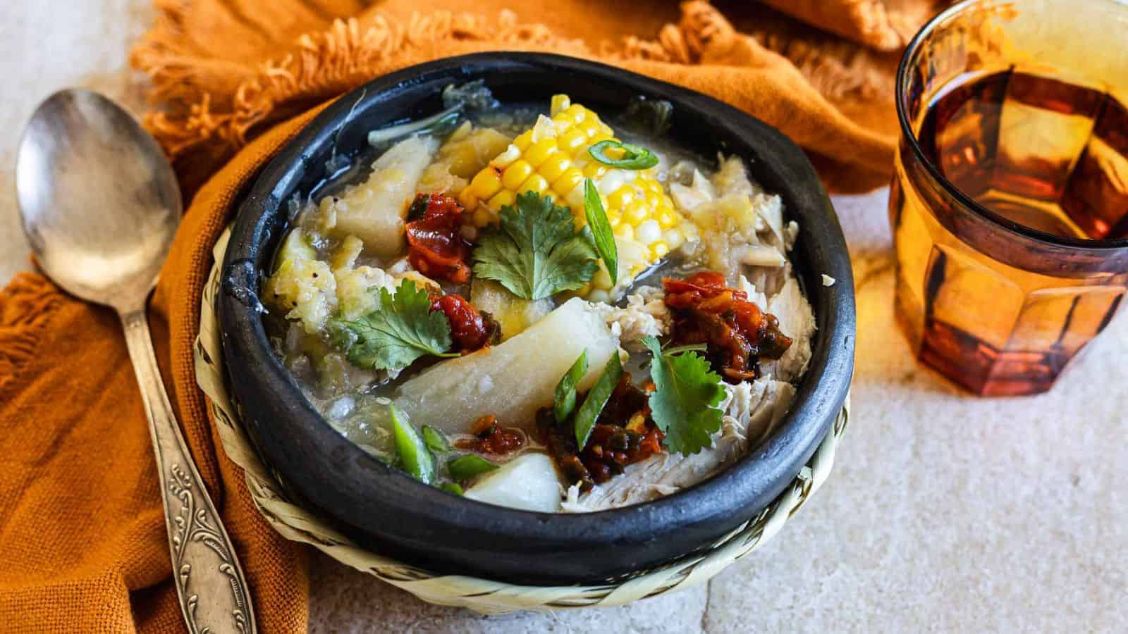 A bowl of hearty soup with corn, meat, potatoes, and cilantro garnished with chili, placed on a woven mat. A spoon rests beside the bowl on an orange cloth. A glass of amber liquid is to the right.