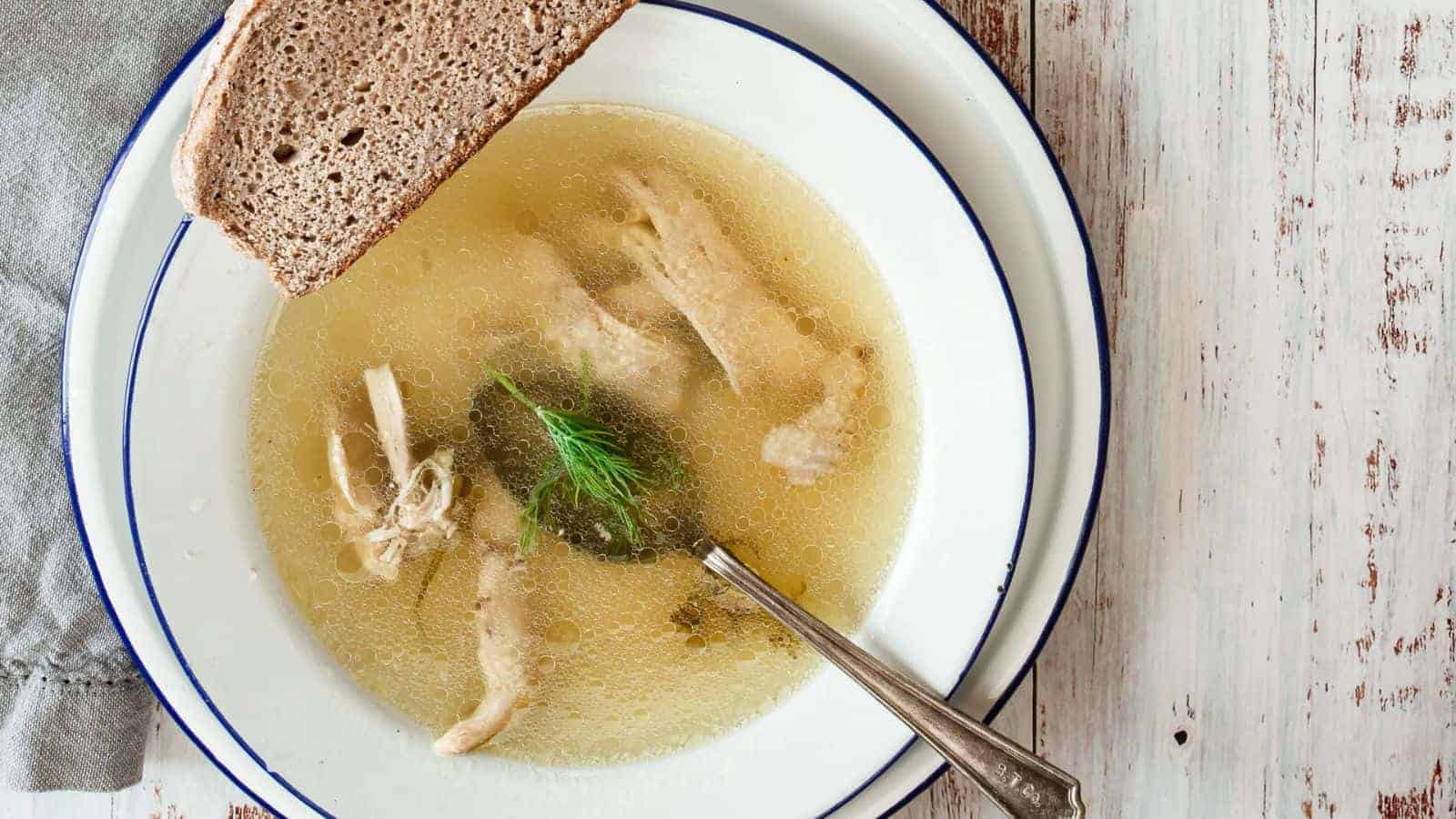 A bowl of clear chicken soup with visible slices of chicken and a sprig of dill. A slice of brown bread rests on the rim of the bowl, and a spoon is placed inside. The bowl is set on a wooden table with a cloth napkin beside it.