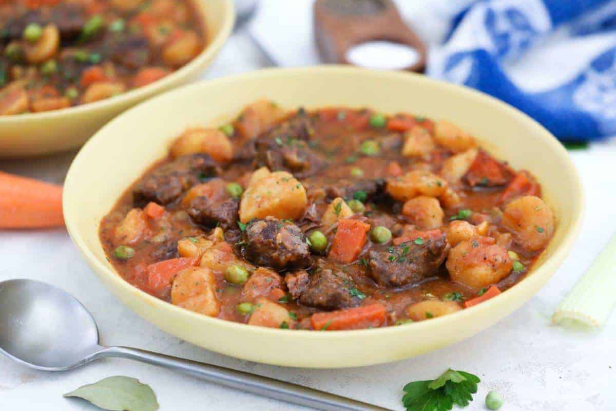 A bowl of beef stew containing chunks of beef, potatoes, carrots, and peas in a thick broth. A spoon lies beside the bowl on a light surface, and a blue and white cloth is partially visible in the background.
