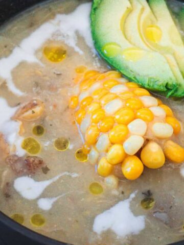 A bowl of creamy soup topped with a piece of corn on the cob, sliced avocado, and a drizzle of cream. A spoon is placed beside the bowl on a wooden surface.