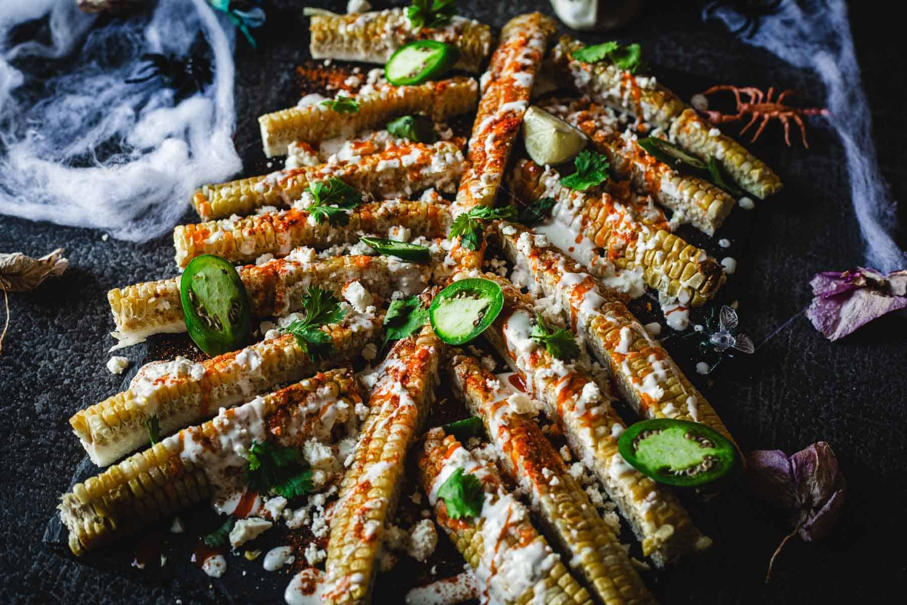 Grilled corn on the cob sliced into halves, arranged in a triangular pattern on a dark surface, topped with creamy sauce, chili powder, cilantro, and lime wedges; garnished with jalapeño slices. Decorative cobwebs and plastic insects surround the dish.