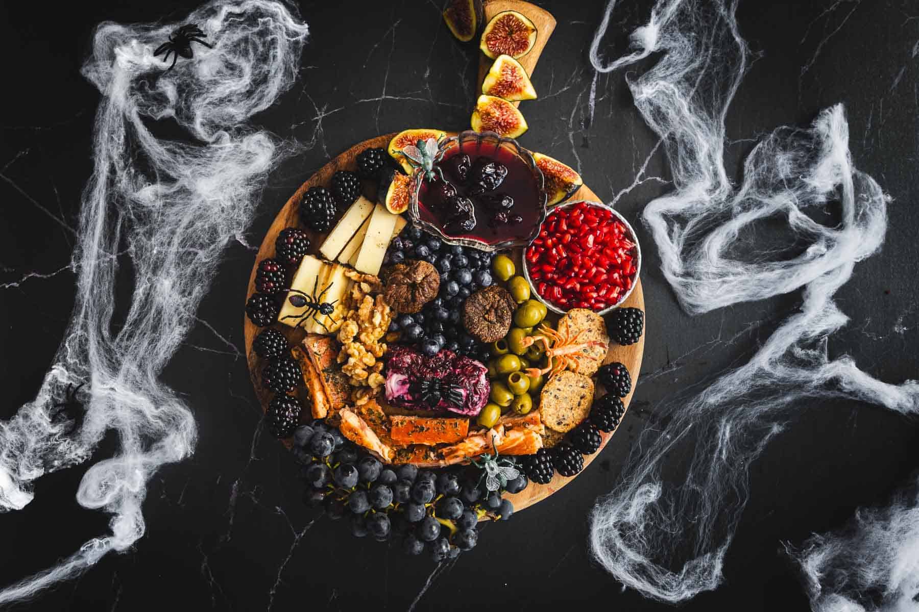 A round wooden board displays an assortment of cheeses, blackberries, figs, pomegranate seeds, grapes, olives, nuts, and crackers. Decorative cobwebs surround the board on a dark surface.