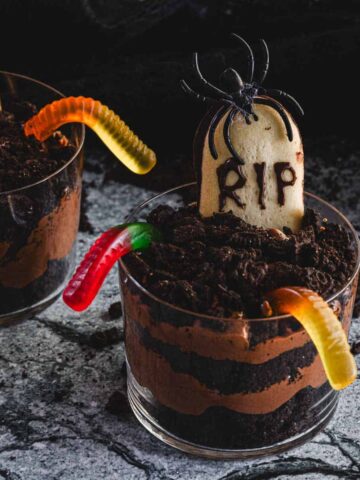 Three cups of layered chocolate pudding topped with crushed cookies and colorful gummy worms are shown. Each cup has a cookie decorated as a tombstone with "RIP" written on it, and a plastic spider is placed on one of the tombstones.