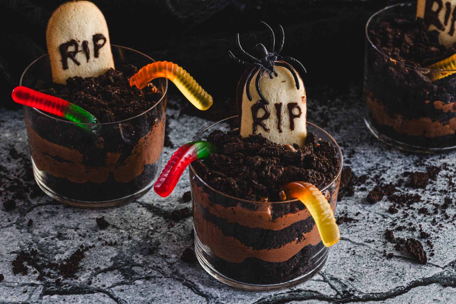 Three cups of layered chocolate pudding topped with crushed cookies and colorful gummy worms are shown. Each cup has a cookie decorated as a tombstone with "RIP" written on it, and a plastic spider is placed on one of the tombstones.
