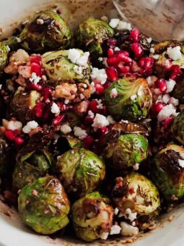 A bowl of roasted Brussels sprouts topped with crumbled feta cheese, pomegranate seeds, and a drizzle of balsamic glaze. A spoon rests on the edge of the bowl, which sits on a light wooden surface.