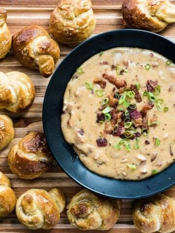 Bacon beer cheese in a dark bowl surrounded by pretzel knots on a cutting board.