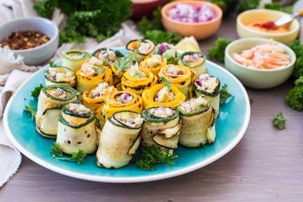A plate of rolled zucchini and squash slices filled with a mixture of shrimp, diced red onion, and herbs. The rolls are garnished with parsley. In the background, there are small bowls containing additional ingredients like shrimp and spices.